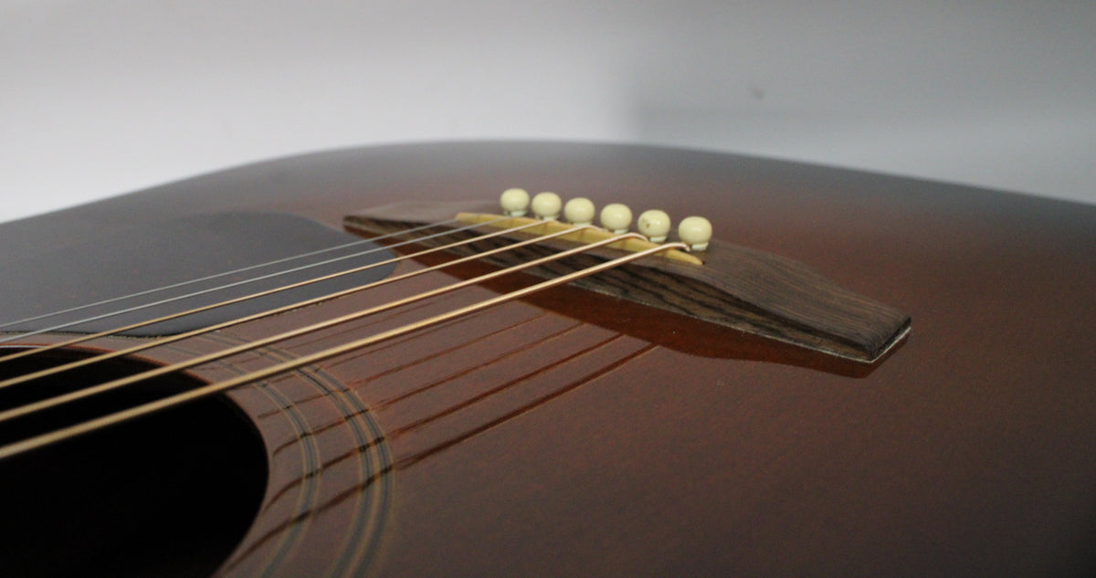 Close-up of the sunburst acoustic guitar's lower bout, showing the dark wood bridge, six cream bridge pins, and light-colored strings.