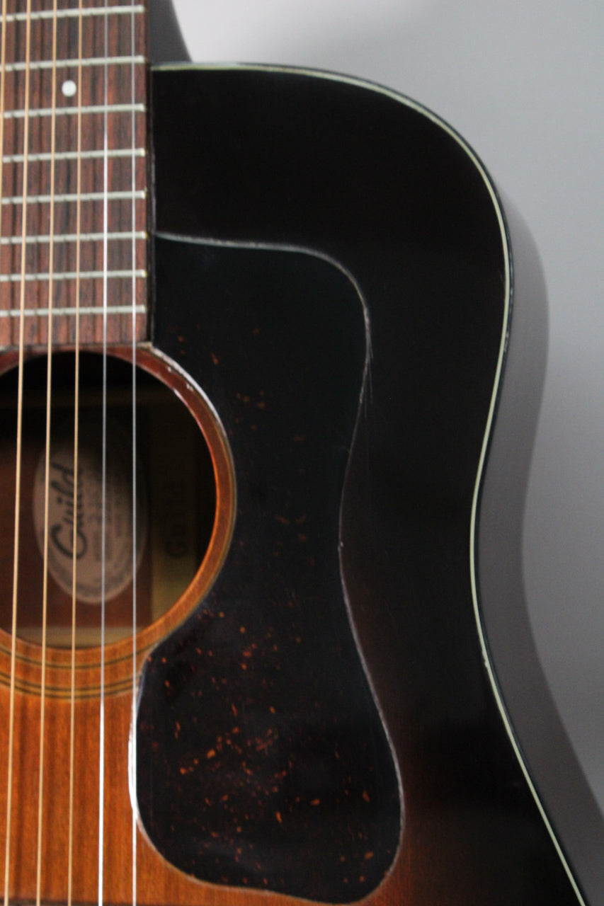 The upper bout of a sunburst acoustic guitar, showing the dark brown pickguard with orange flecks and finish checking.