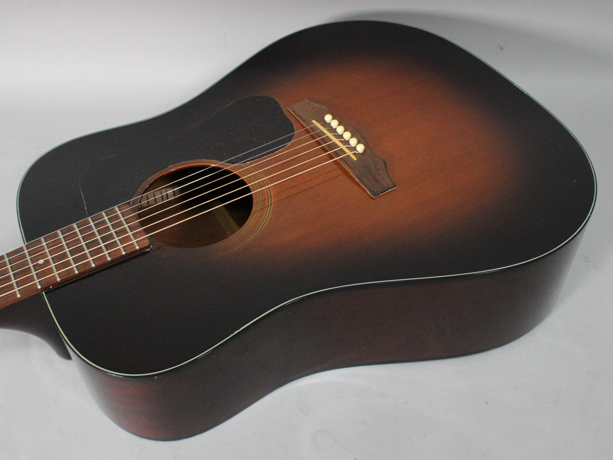 The sunburst acoustic guitar is angled down and to the right, showing its dark brown body, pickguard, and light wear.
