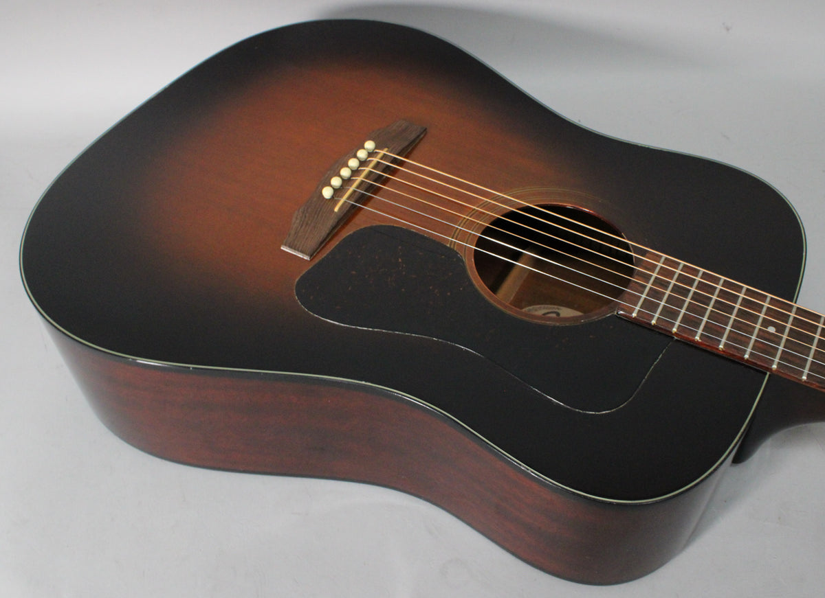 The sunburst acoustic guitar is angled down and right, showing its dark brown top, black pickguard, and mahogany sides.