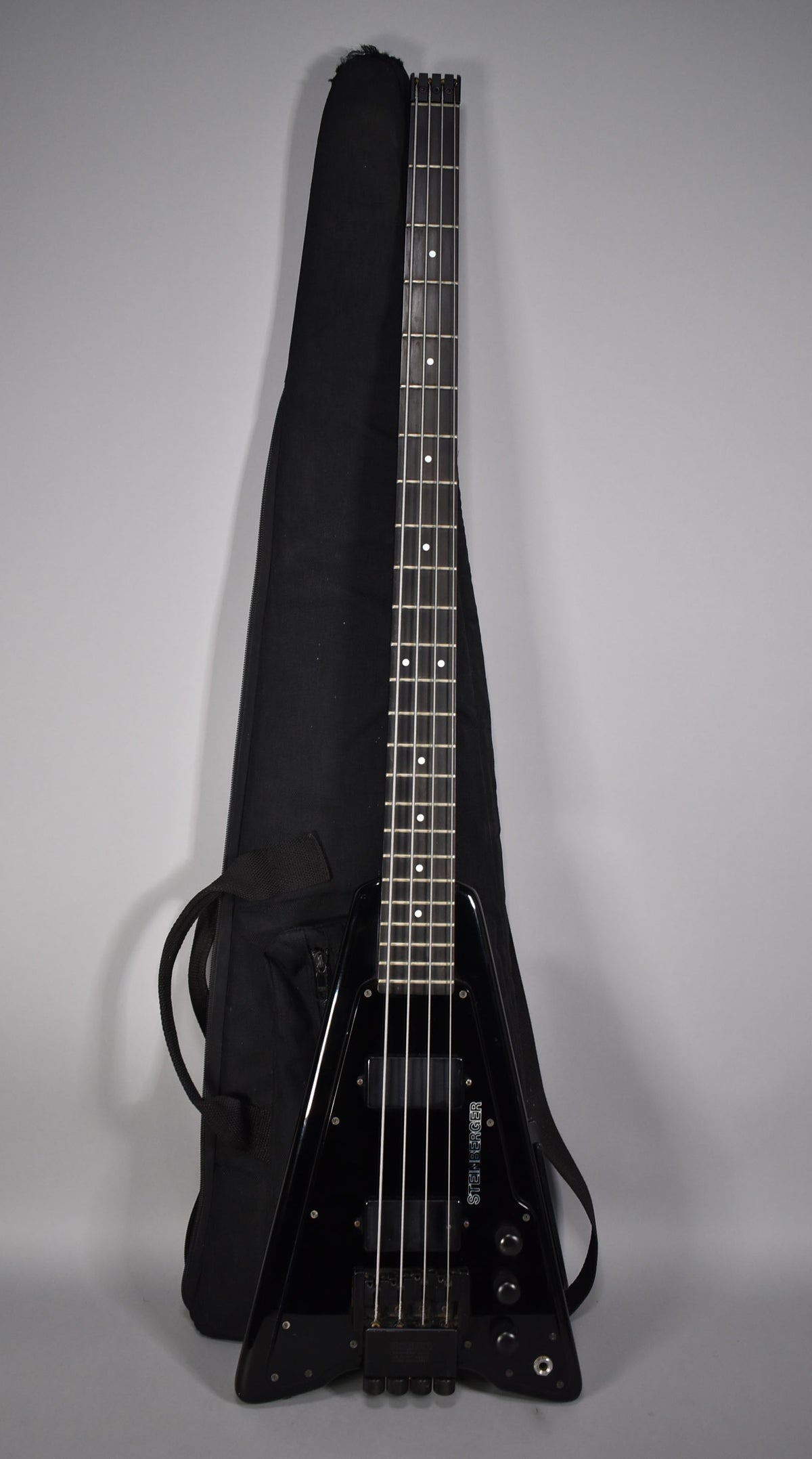 Full-length view of a black V-shaped bass guitar with a headless neck, next to its black gig bag.