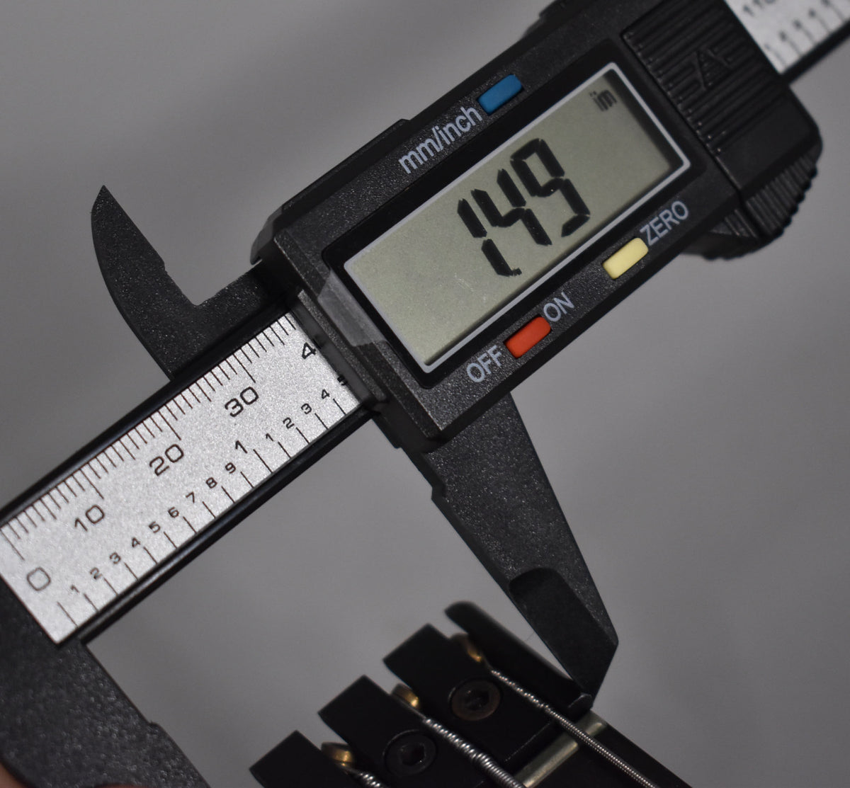 A digital caliper measures the black headstock of the bass, showing 1.49 inches on its display.