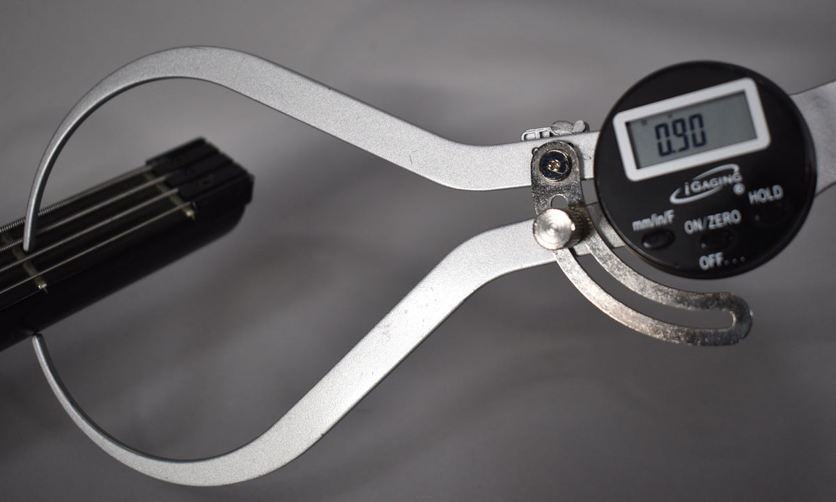 A digital caliper measures the black bass guitar's fretboard and strings, displaying "0.90" on its screen.