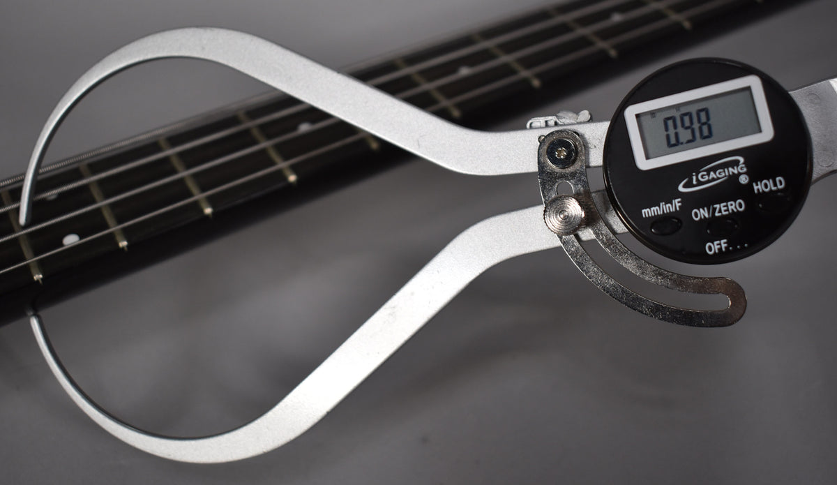 A digital caliper measures the string height at the 12th fret of the bass, showing a reading of 0.98.