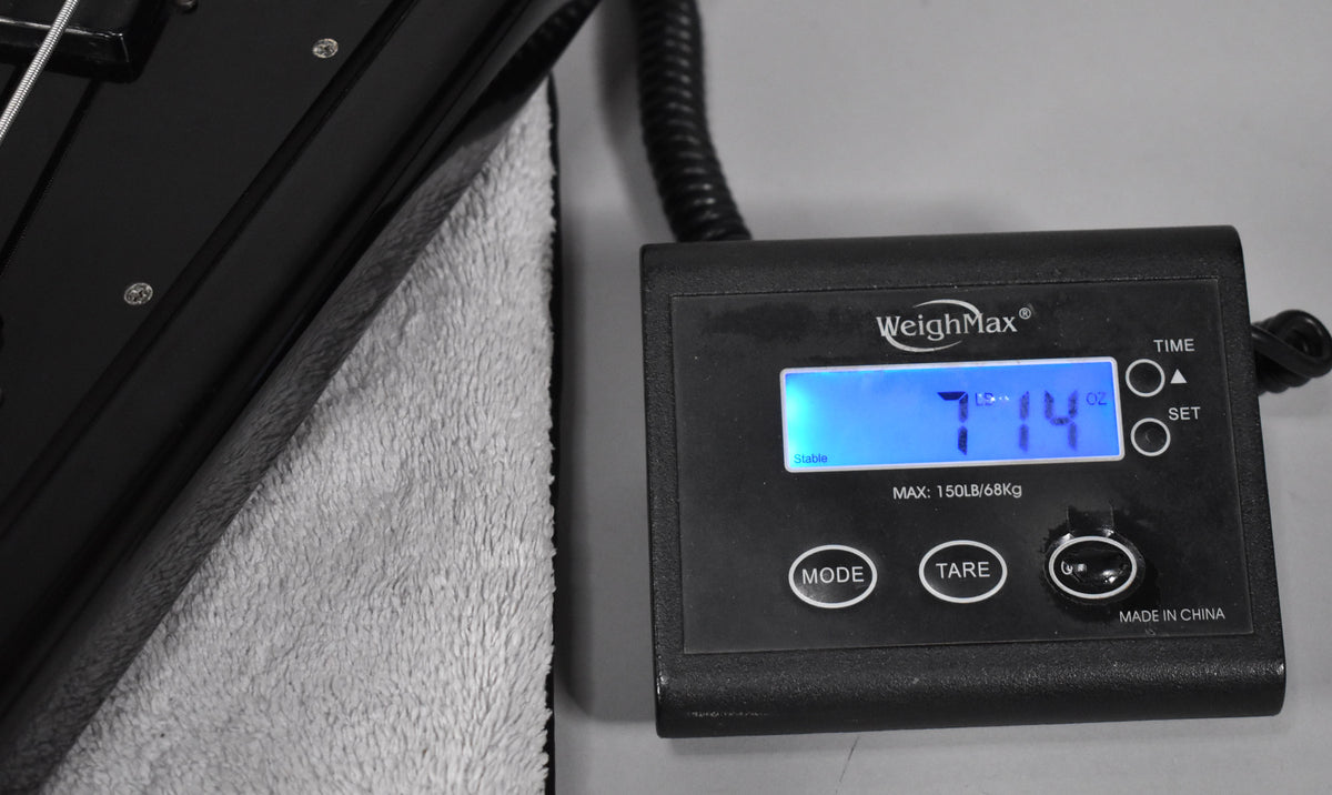 The black bass guitar's body rests on a towel, with a digital scale displaying "7.14 lbs" to the right.