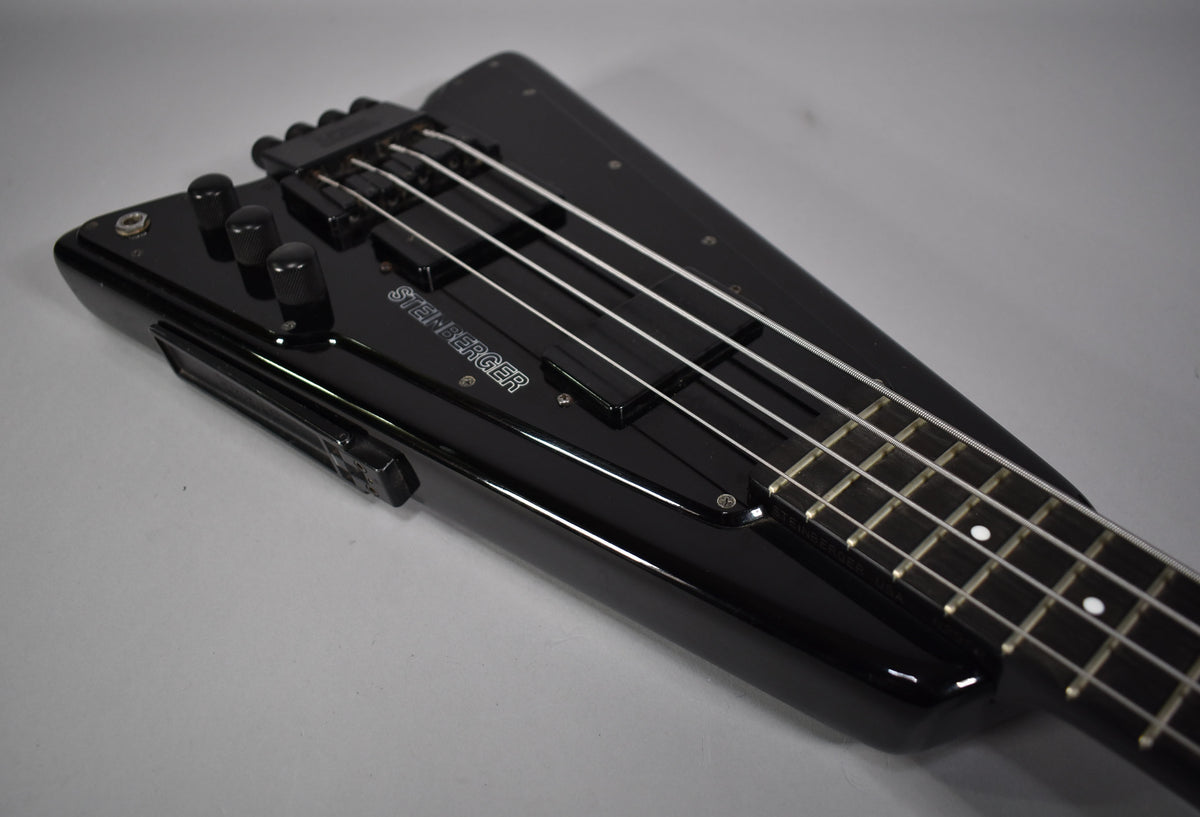 The black headless bass is angled from the upper left, showing its glossy body, two pickups, three control knobs, and part of the fretboard.
