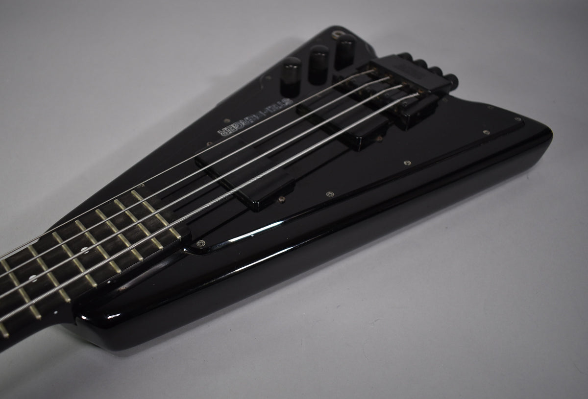 Close-up of the glossy black bass body, angled from the neck, showing two pickups, three control knobs, and string tuners.