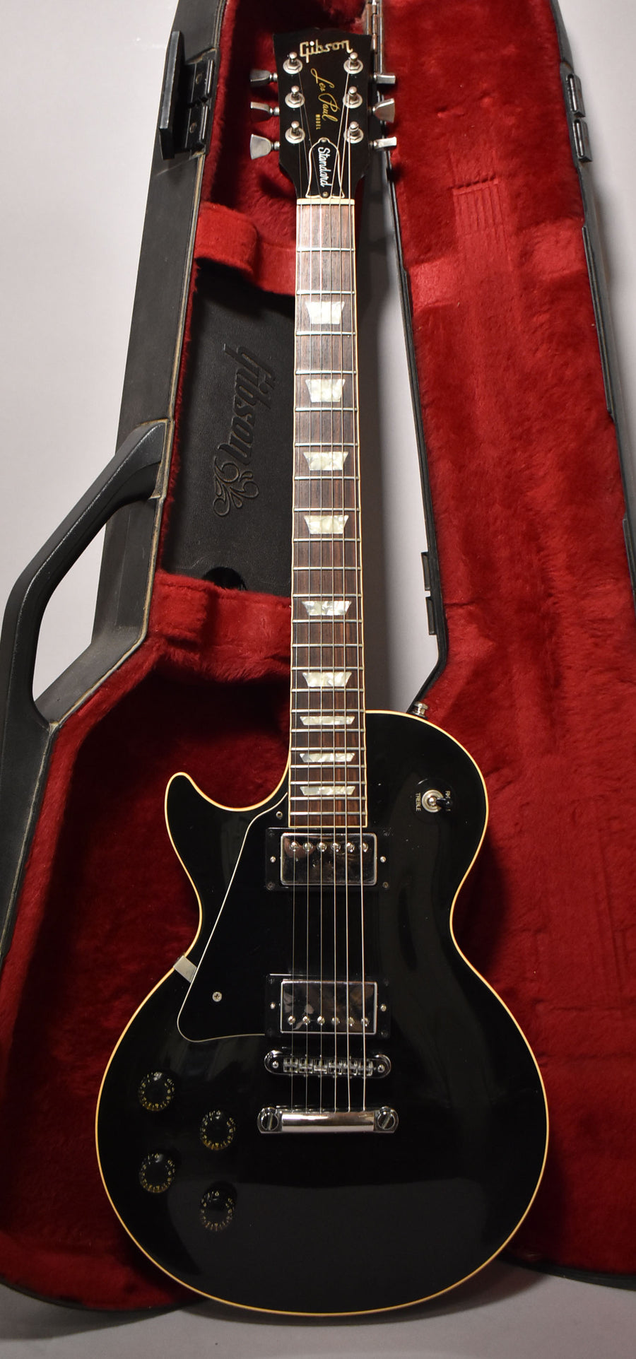 Full-length view of a glossy black Les Paul with cream binding, chrome hardware, trapezoid inlays, and a red-lined hard case.