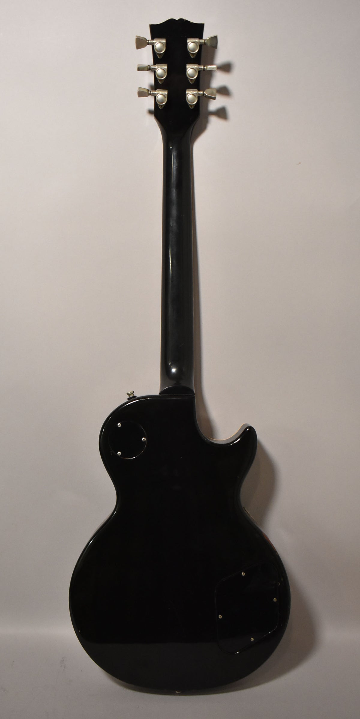 Full rear view of a glossy black Les Paul, showing the headstock with six tuners, neck, and body with control cavity covers.