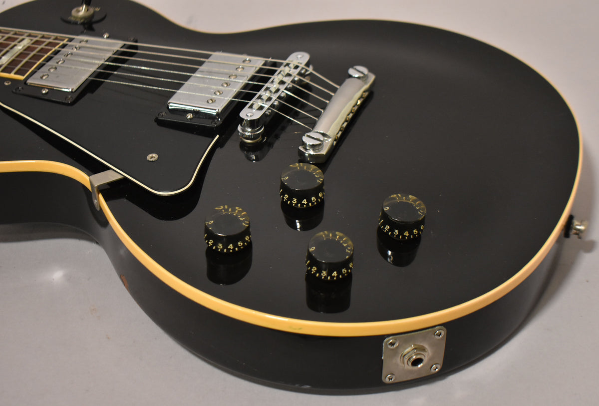 Close-up of a black lefty electric guitar body with cream binding, showing four numbered black control knobs and a chrome output jack.