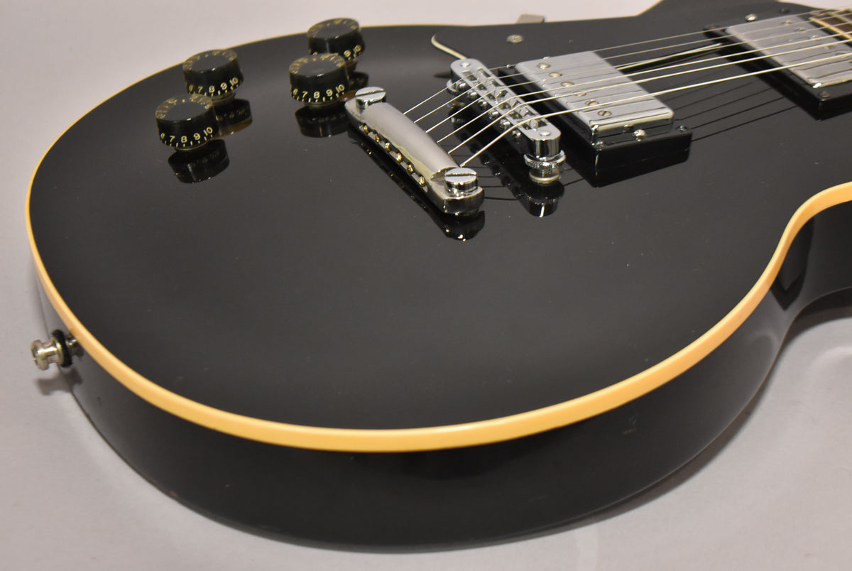 Close-up of a glossy black electric guitar body, showing the chrome bridge, tailpiece, and four black control knobs with white numbers.