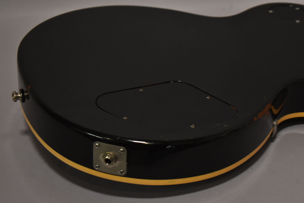 The back of a glossy black guitar, showing the control cavity cover, output jack, and a strap button, with some edge wear.