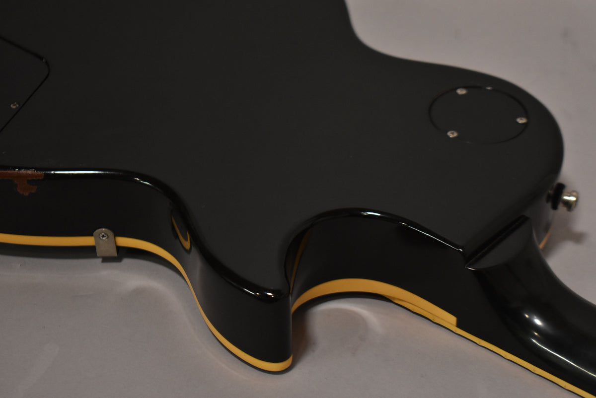 The back of a glossy black guitar body with cream binding, showing a control cavity cover and strap button, with wear on the upper edge.