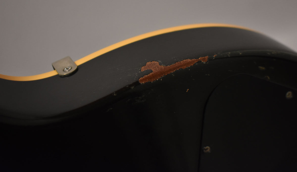 Close-up of a black guitar body with cream binding, showing a large chip in the finish revealing reddish wood, and a strap button.