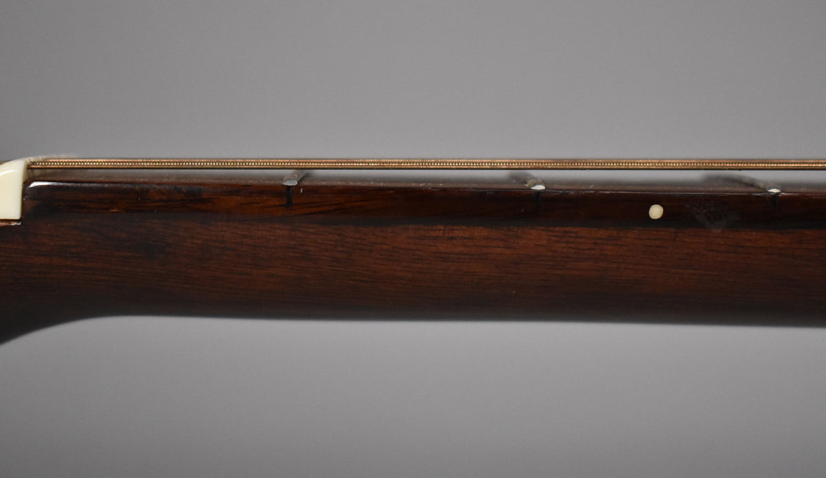 Close-up of the dark brown neck and fretboard from the side, showing the nut, strings, and a single white dot inlay.