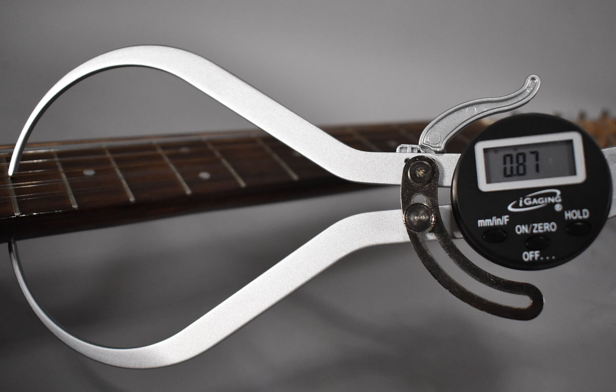 A digital caliper measures the dark wood fretboard, showing 0.87 on its black display.