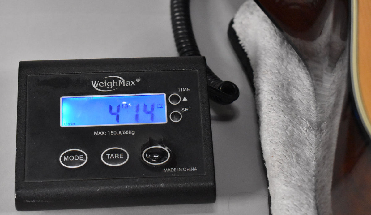A black digital scale displays "4 LB 14 OZ" next to the guitar's dark brown body, resting on a white plush lining.