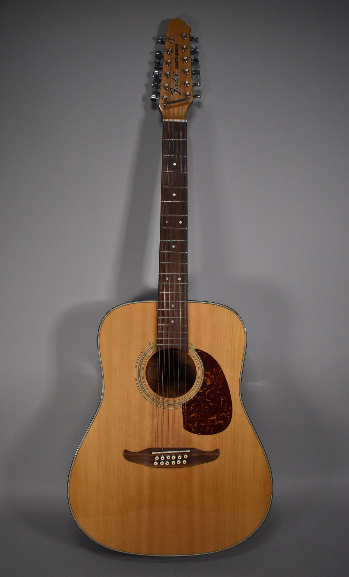 Full front view of a natural finish 12-string acoustic guitar with a dark tortoiseshell pickguard and a unique wavy bridge.