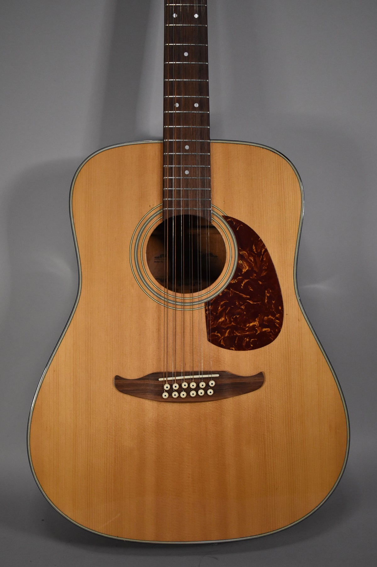 Full front view of a natural finish 12-string acoustic guitar with a dark tortoiseshell pickguard and a dark wood bridge with 12 white pins.