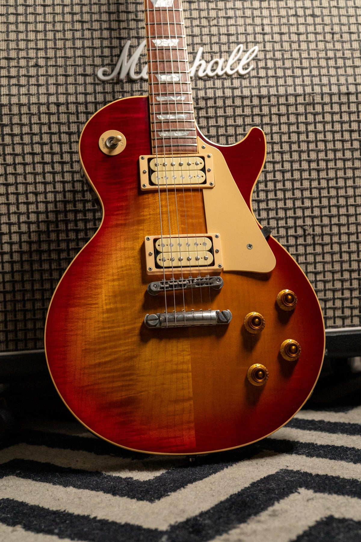 Full-body view of a cherry sunburst guitar with a flame top, cream humbuckers, and gold top hat knobs, leaning against an amp.