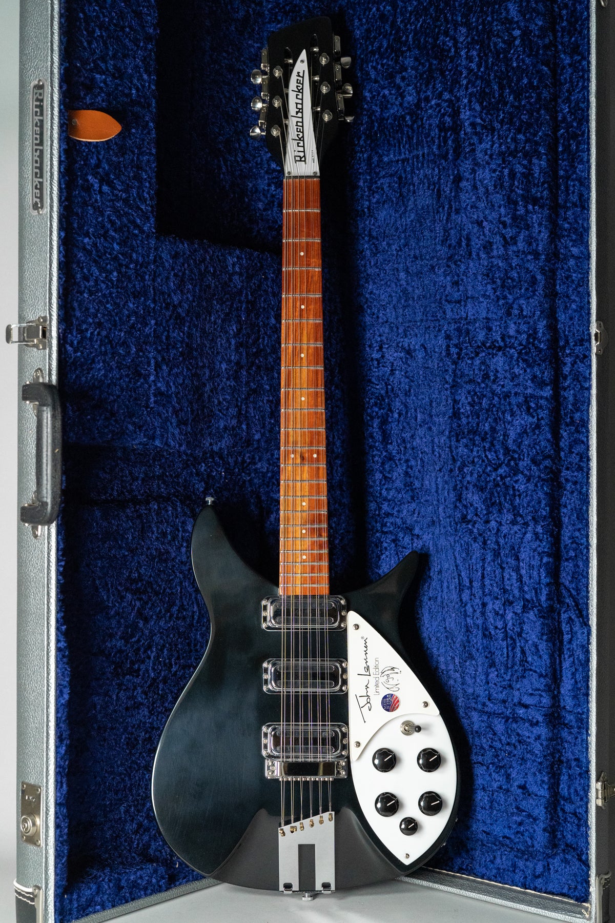A black 12-string guitar with a white pickguard, chrome hardware, and a dark wood fretboard, resting in a blue velvet case.