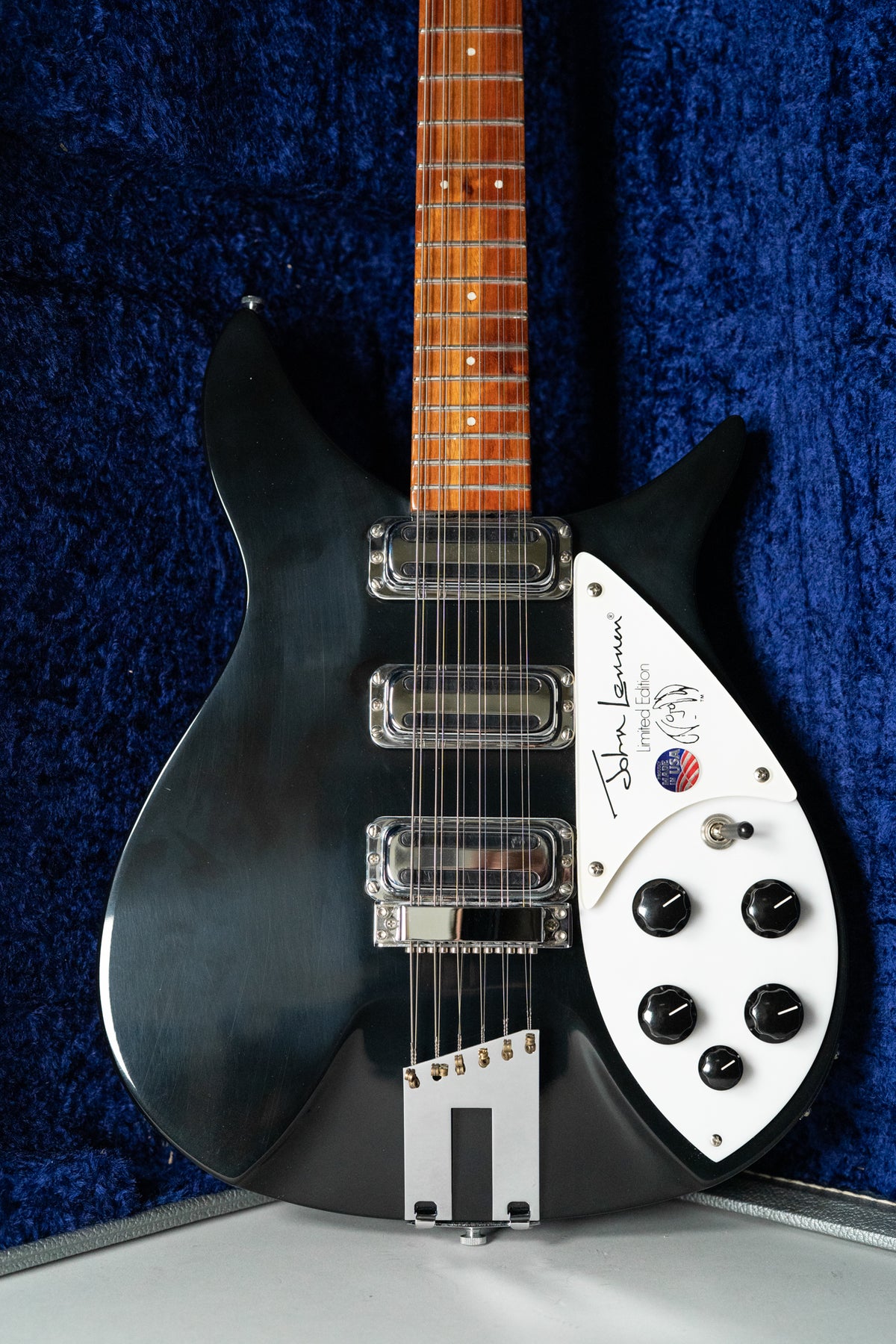 The black 12-string guitar is framed from the upper body to the bridge, showing three chrome pickups and a white pickguard with five black knobs.