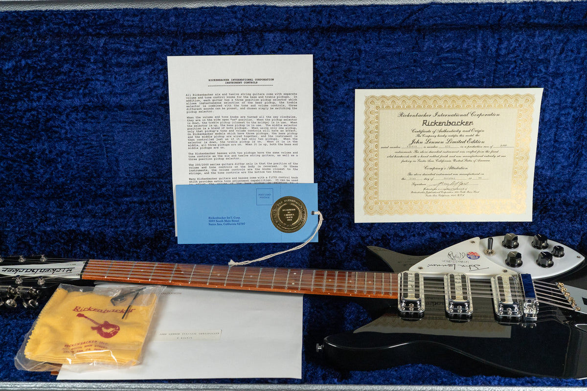 Overhead view of a black 12-string guitar in a blue velvet case, with a certificate, manual, and yellow polishing cloth.