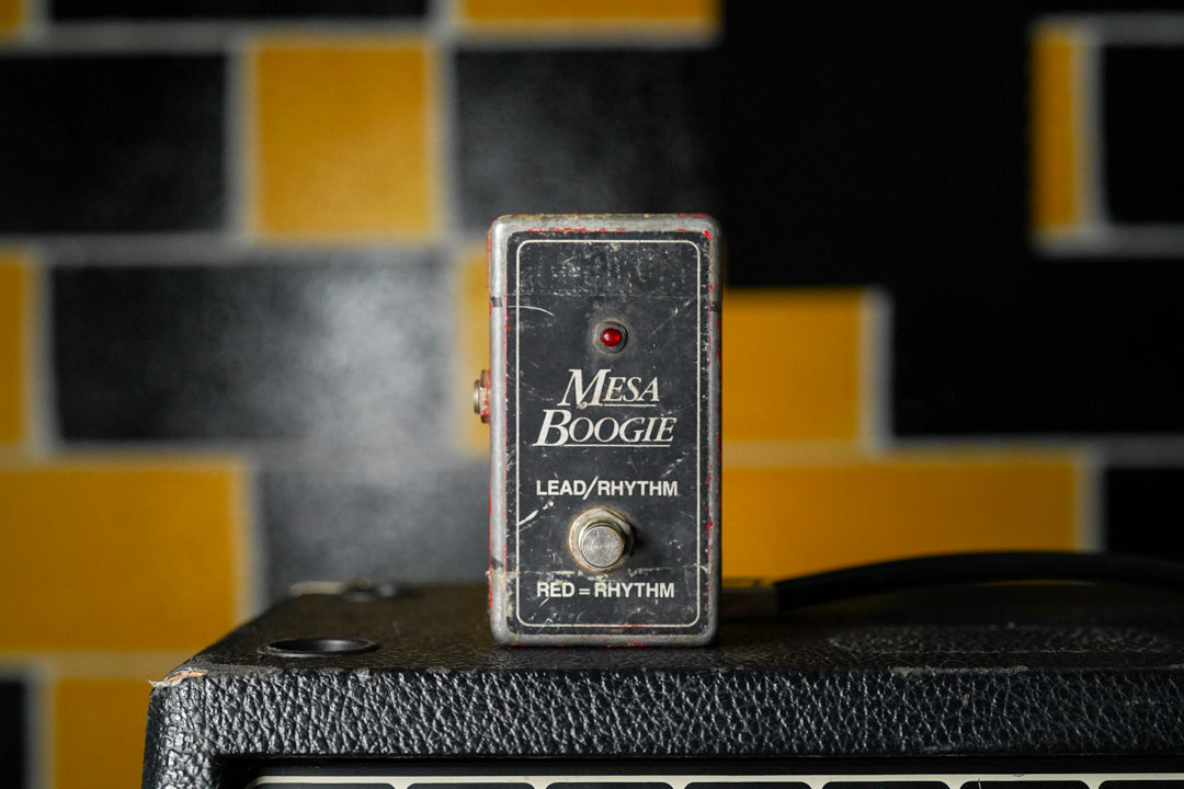 A worn, rectangular black footswitch with a red LED and a single silver button, sitting on top of a black textured amplifier.
