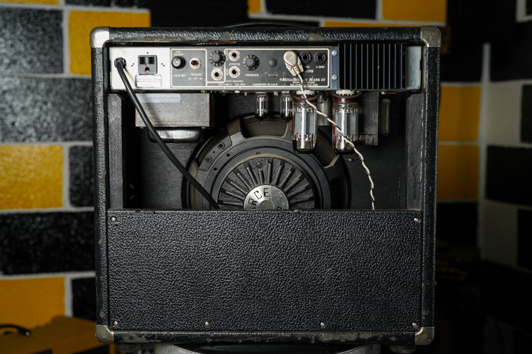 Rear view of a black textured combo amp, showing the speaker, four glowing tubes, and a metal control panel with jacks and knobs.