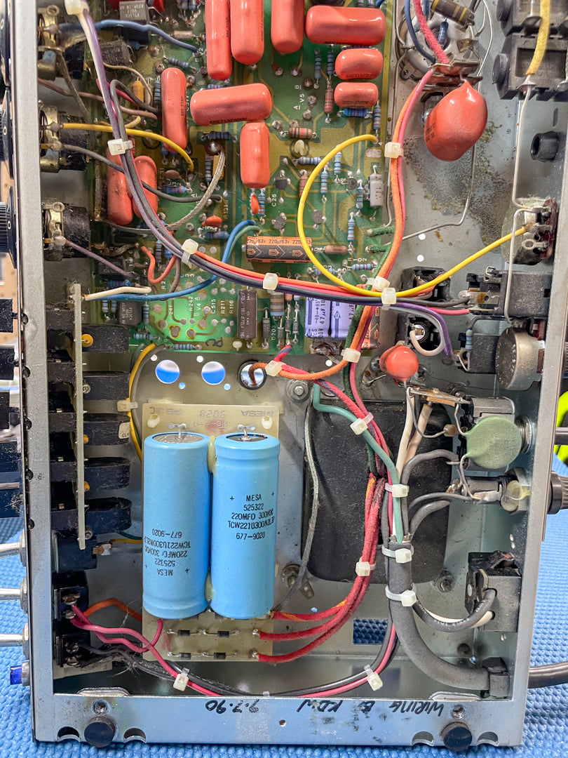 Top-down view of the amp's internal circuitry, showing a green circuit board with orange capacitors, two large blue capacitors, and wiring.