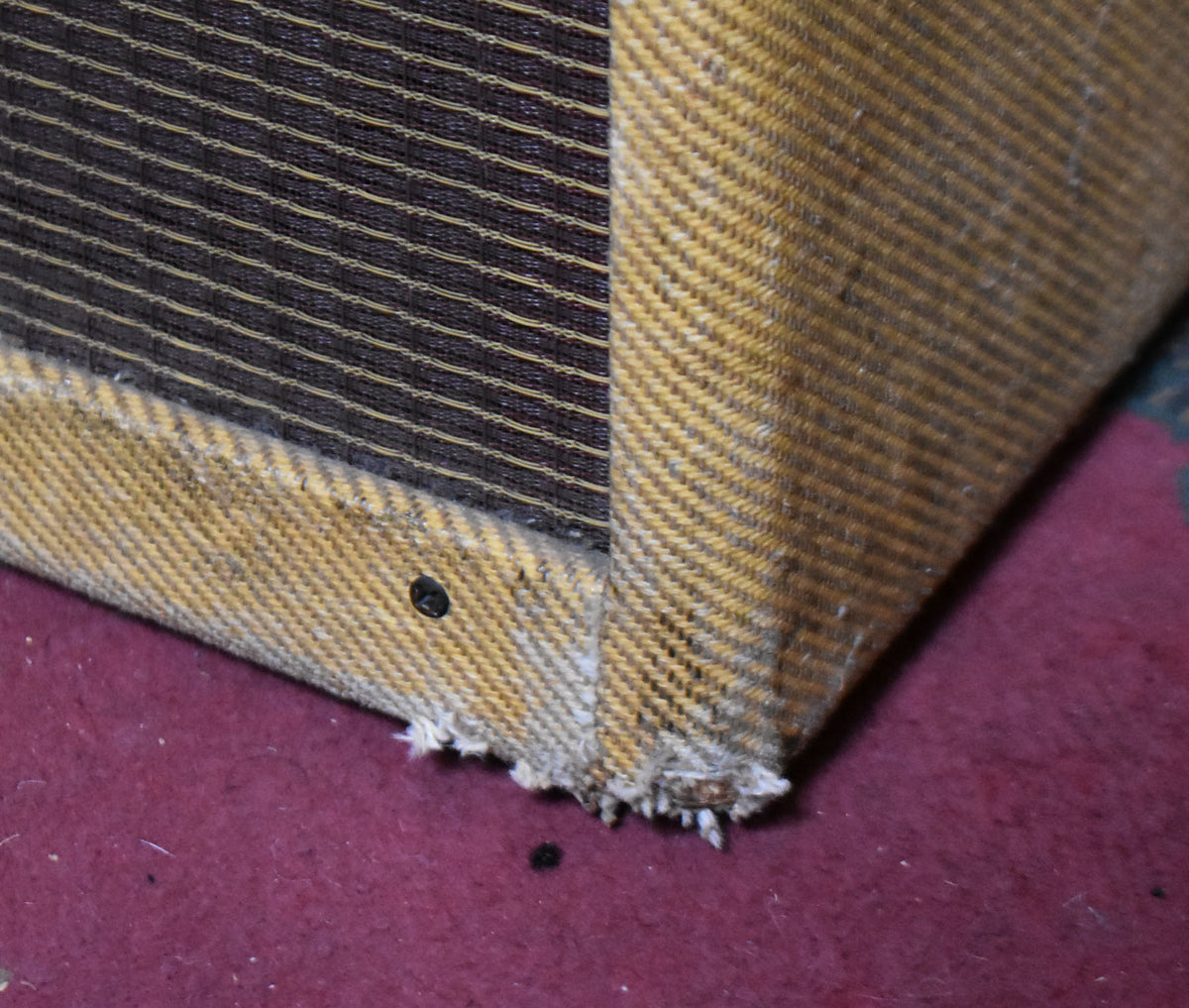 Close-up of the amp's lower left corner, showing frayed tweed fabric, a black screw, and a dark brown grille with horizontal stripes.