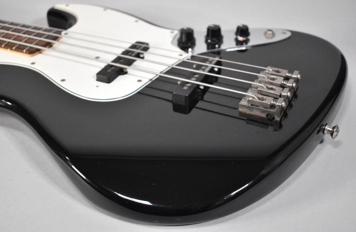 Close-up of a black gloss bass body with a white pickguard, two single-coil pickups, and a chrome bridge with some tarnishing.