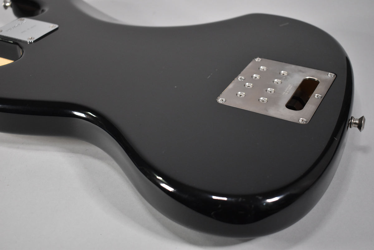 The back of a glossy black bass body, angled from the lower left, shows a chrome neck plate and a rectangular control cavity cover.