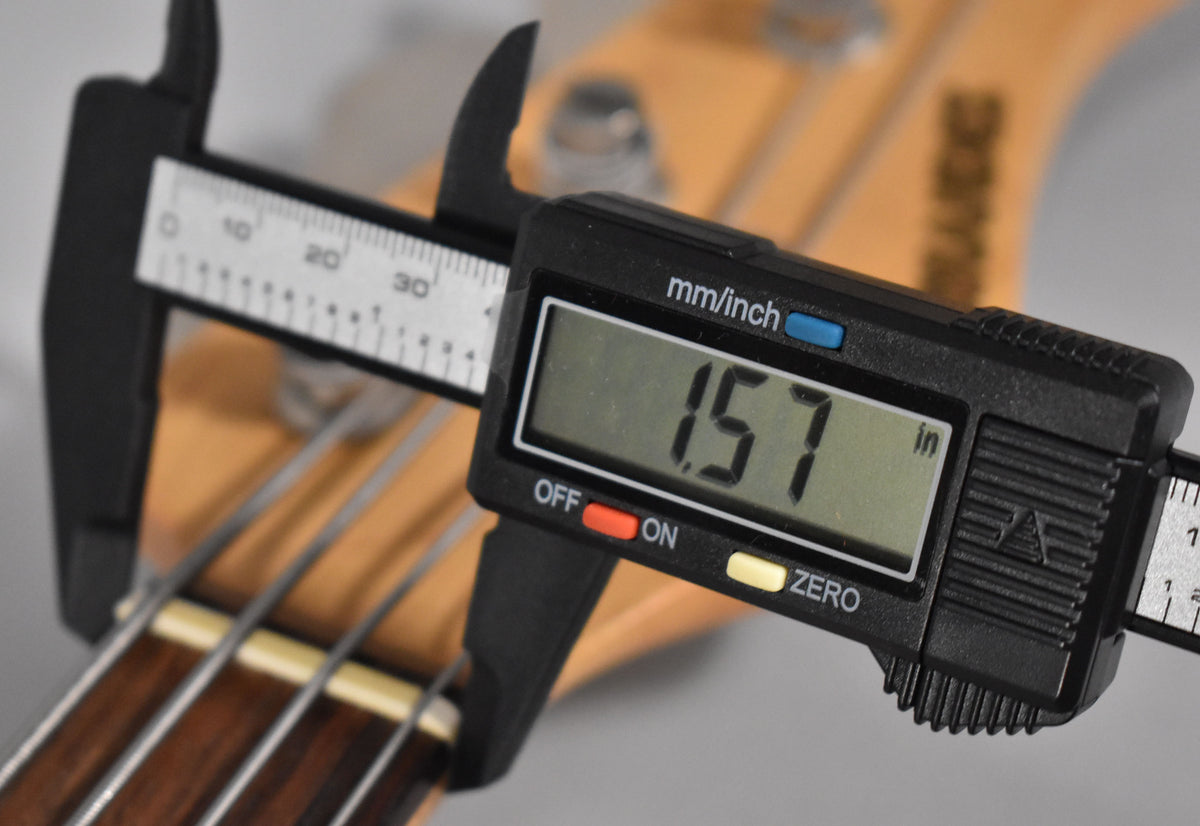 A digital caliper measures the bass's nut width at 1.57 inches, shown against the light brown neck and dark fretboard.
