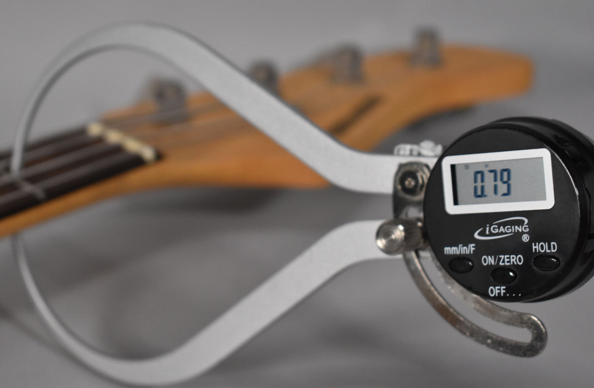 A digital caliper measures the bass's fretboard relief at 0.79, with the light-colored neck and dark fretboard blurred in the background.