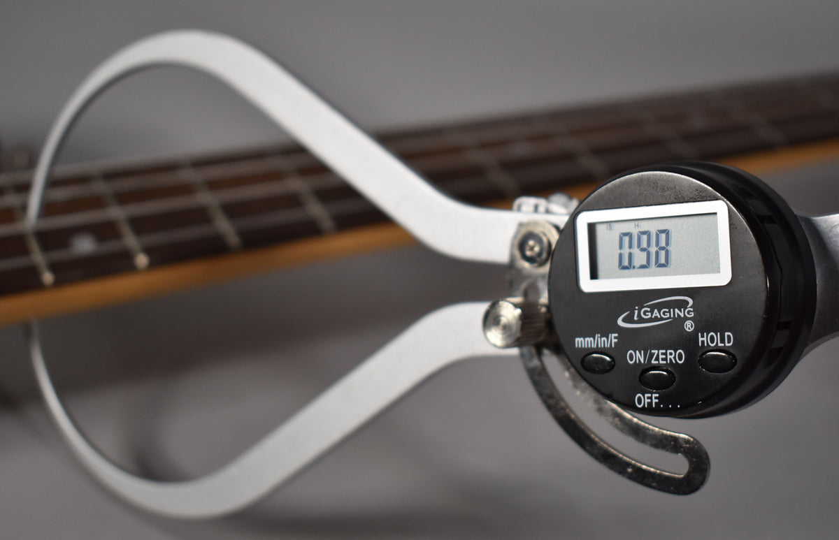 A digital caliper measures the string height of the bass, showing "0.98" on its screen, with the fretboard visible behind it.