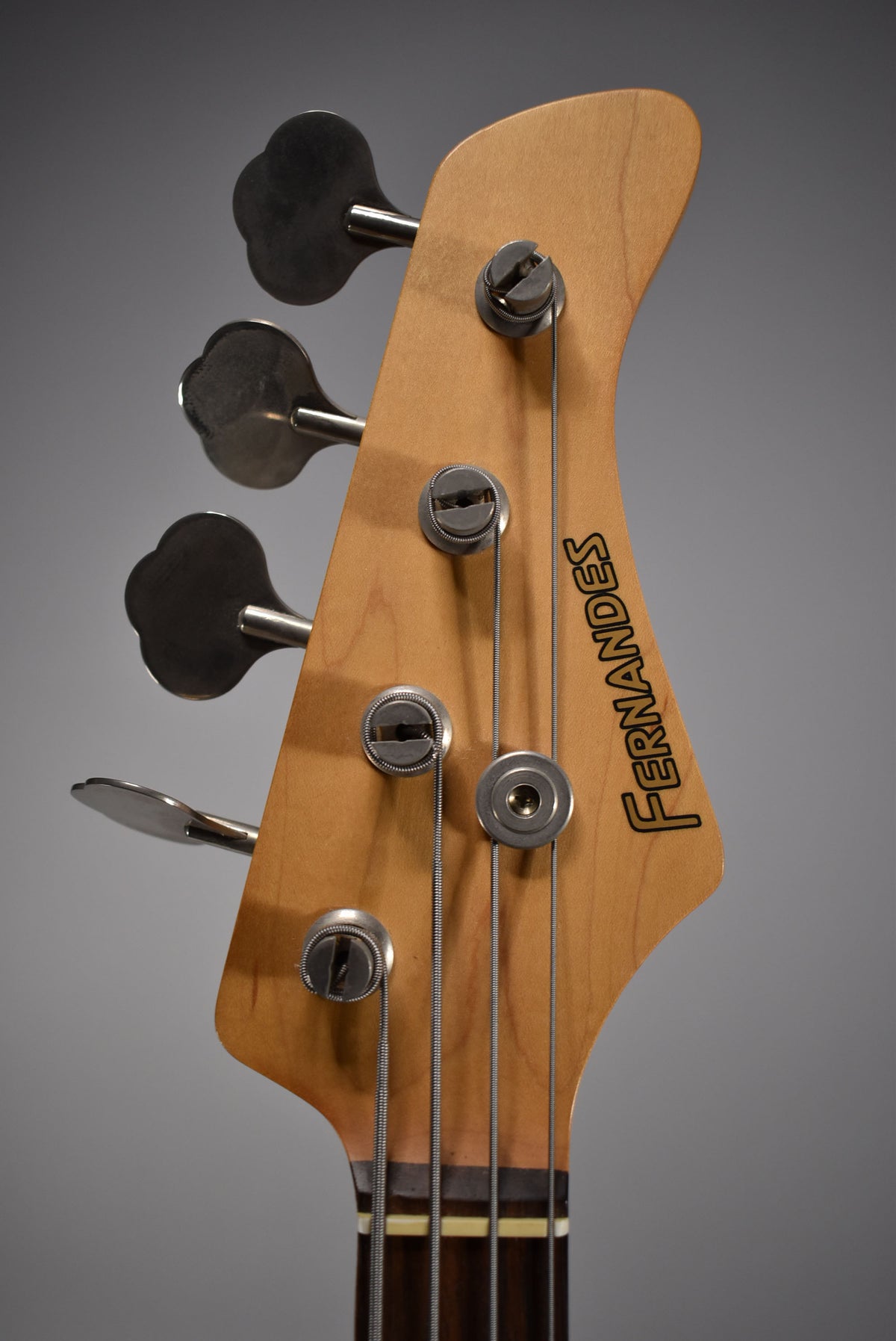 Close-up of the bass headstock, angled left, showing the natural wood finish, four clover-leaf tuners, and string tree.