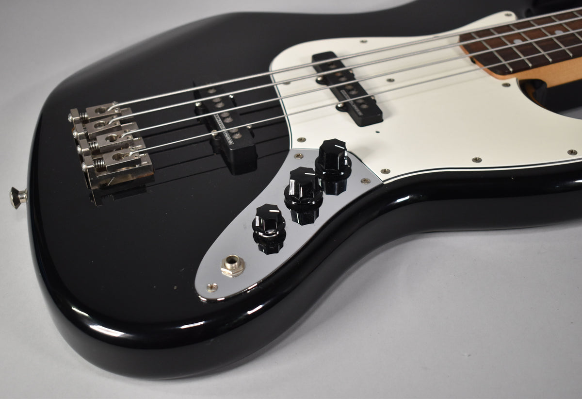 Close-up of a black gloss bass guitar body, showing the chrome bridge, two black pickups, white pickguard, and three black control knobs on a chrome plate.