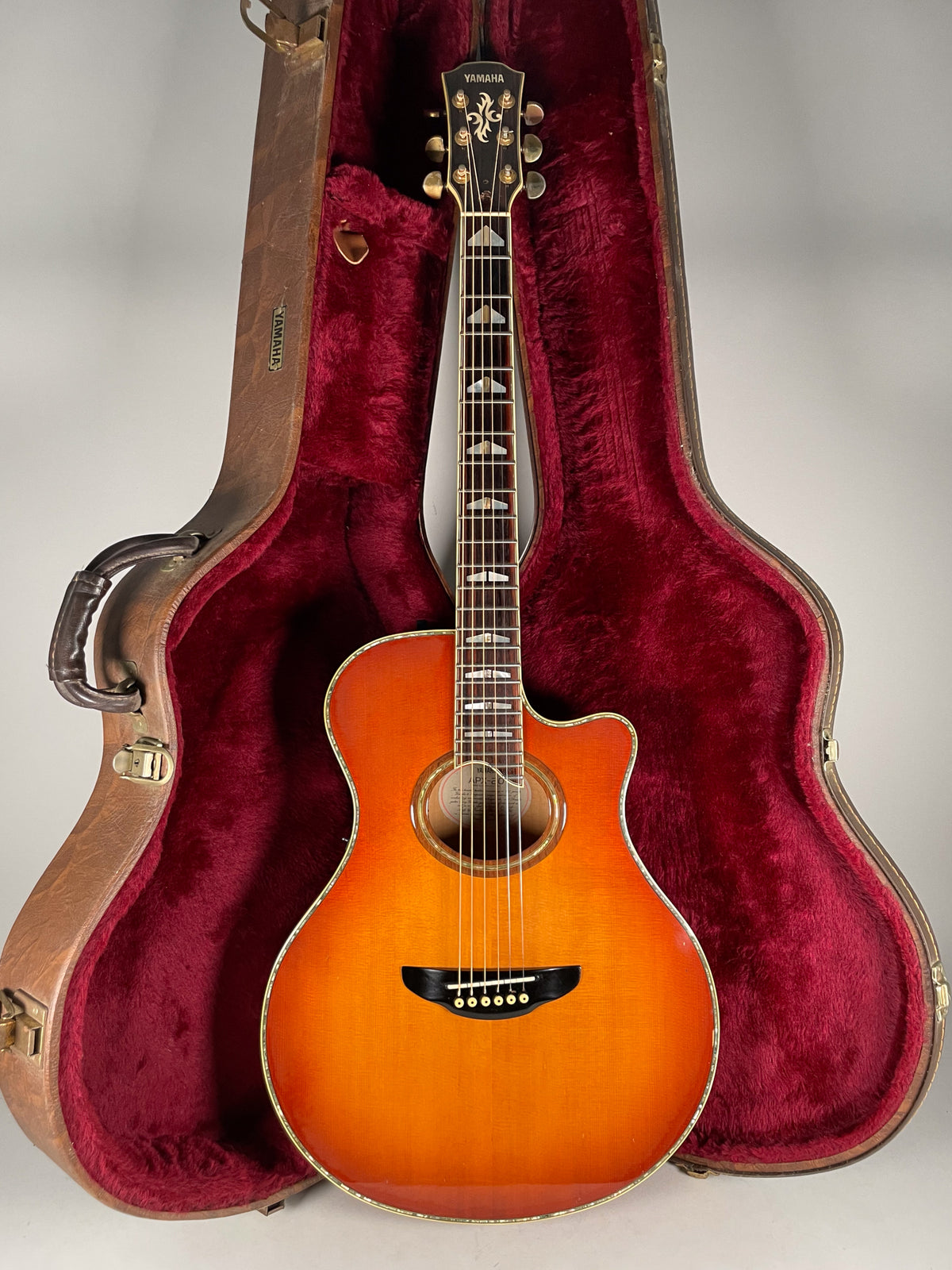Full-length view of a sunburst cutaway acoustic guitar with ornate headstock and triangular fretboard inlays, standing in its open, red-lined hard case.