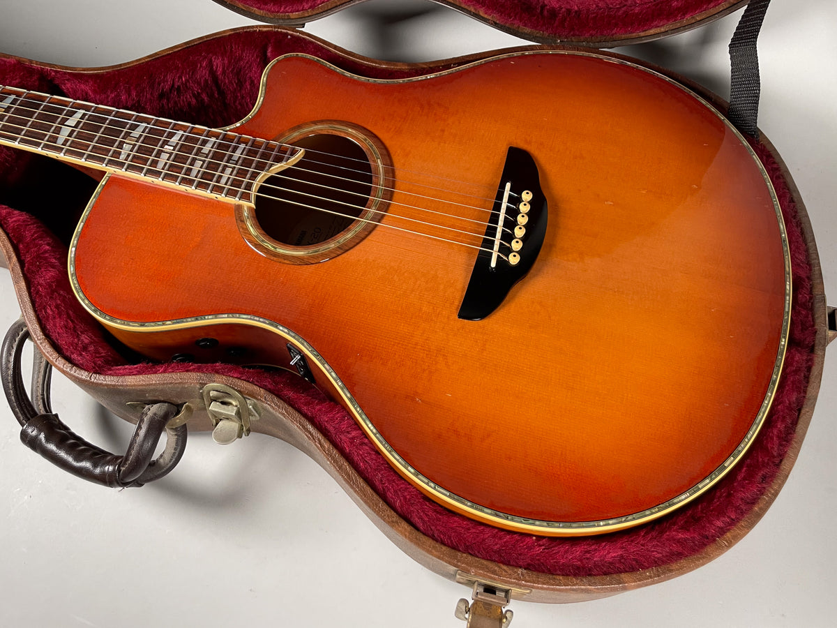Sunburst acoustic guitar with a cutaway body, visible preamp controls, and a black bridge, resting in a red-lined hard case.