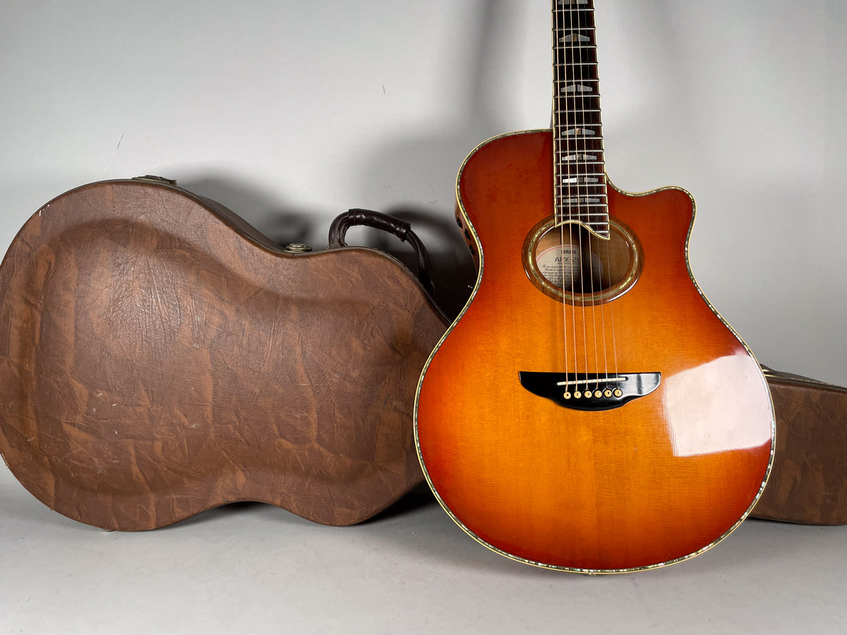 A sunburst acoustic guitar with a cutaway body and black bridge, angled next to a brown, textured hard case.