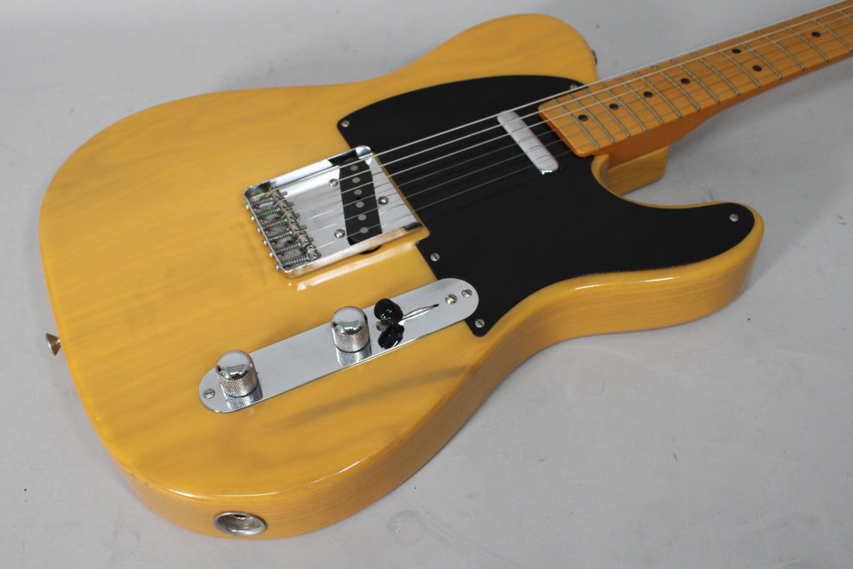 Angled view of a butterscotch blonde Telecaster body with a black pickguard, chrome bridge, control plate, and maple fretboard.