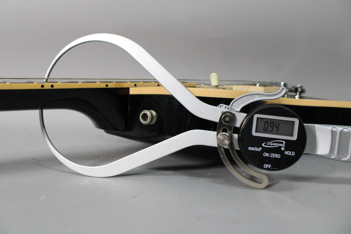 A digital caliper measures the black guitar's neck thickness at 0.94 inches, showing the fretboard and part of the body.