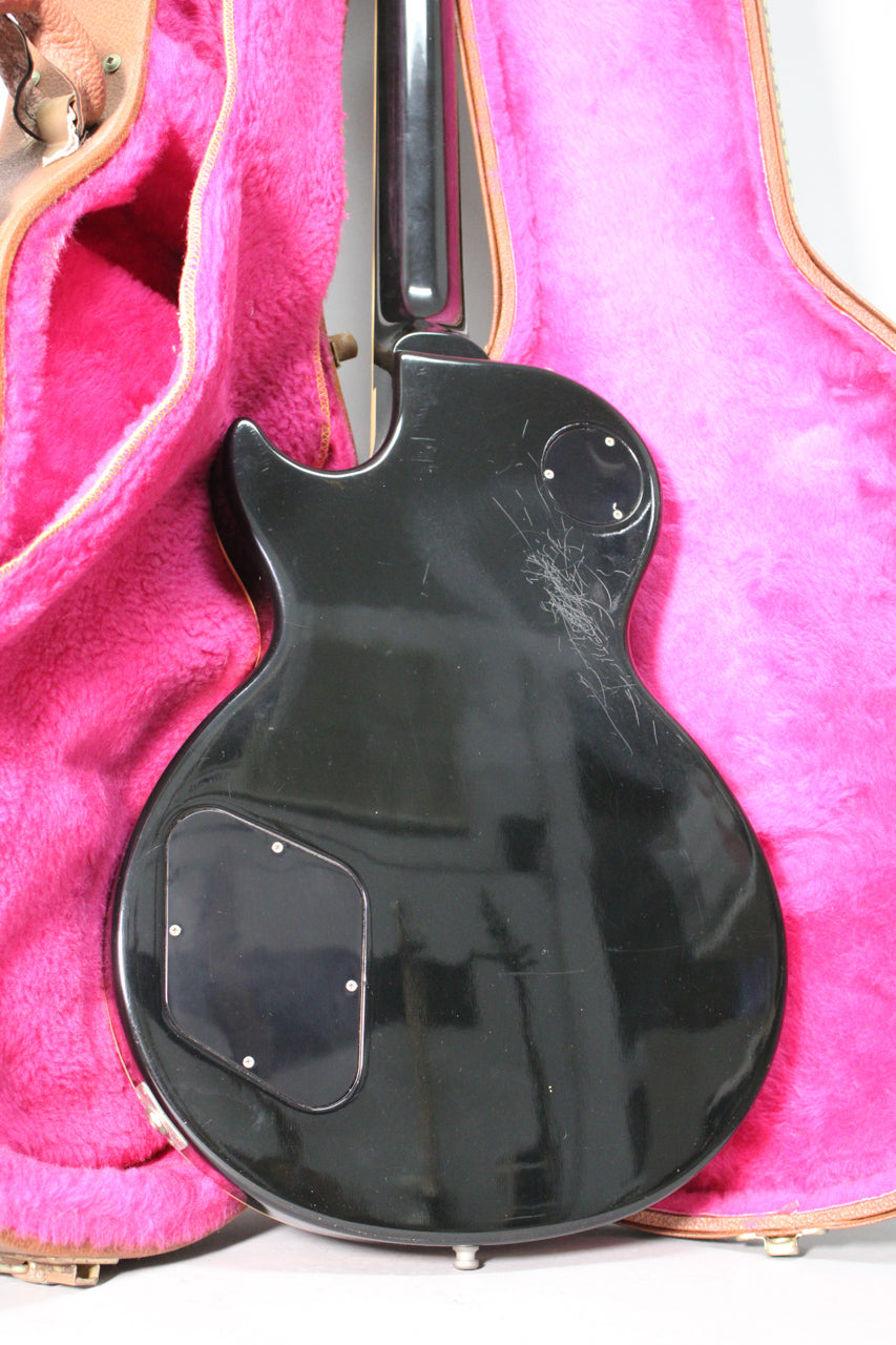 The guitar's glossy black back has many deep scratches, with a control cavity cover and a switch plate, resting in a pink-lined case.