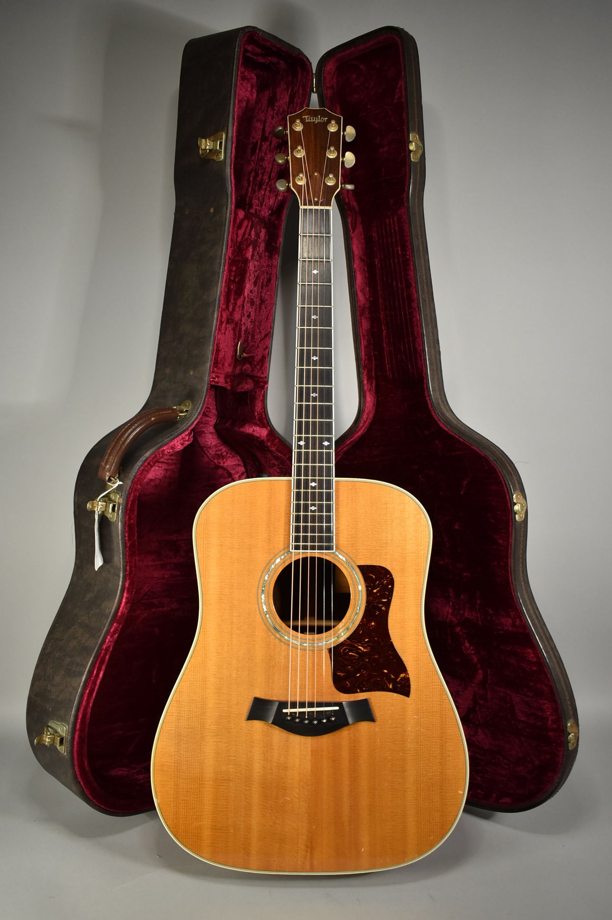 Full-length view of a natural finish acoustic guitar with a tortoiseshell pickguard, standing in its open, red velvet-lined hard case.