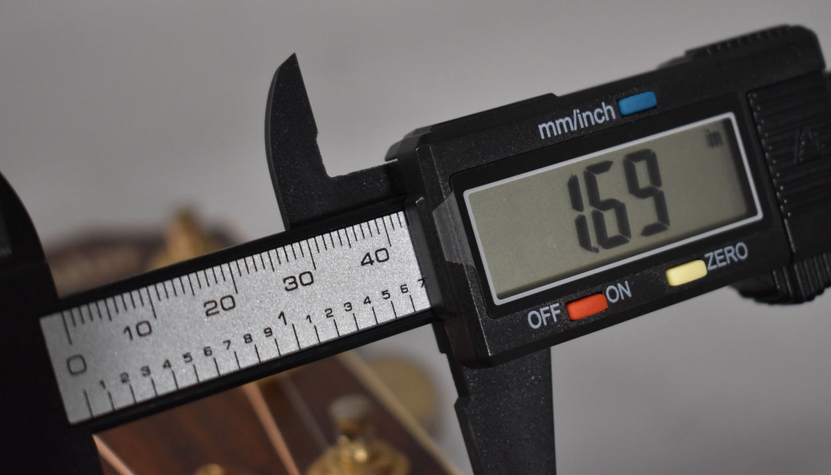 A digital caliper measures the guitar's nut width at 1.69 inches, with the headstock and tuners blurred in the background.