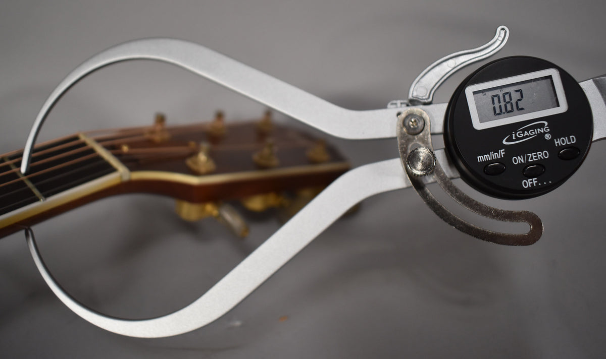 A digital caliper measures the string height at the first fret of the guitar's natural finish neck, showing 0.82mm.