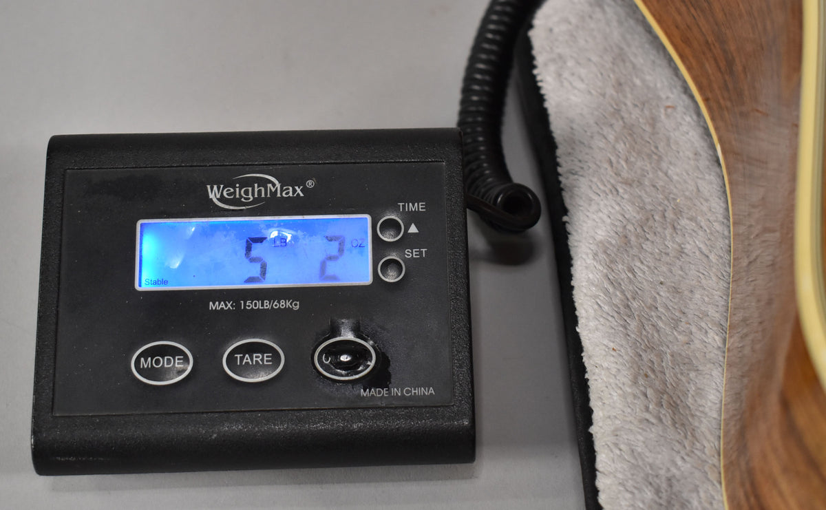 A black digital scale displays "5 LB 2 OZ" next to the guitar's natural wood body and cream binding, resting on a soft grey cloth.