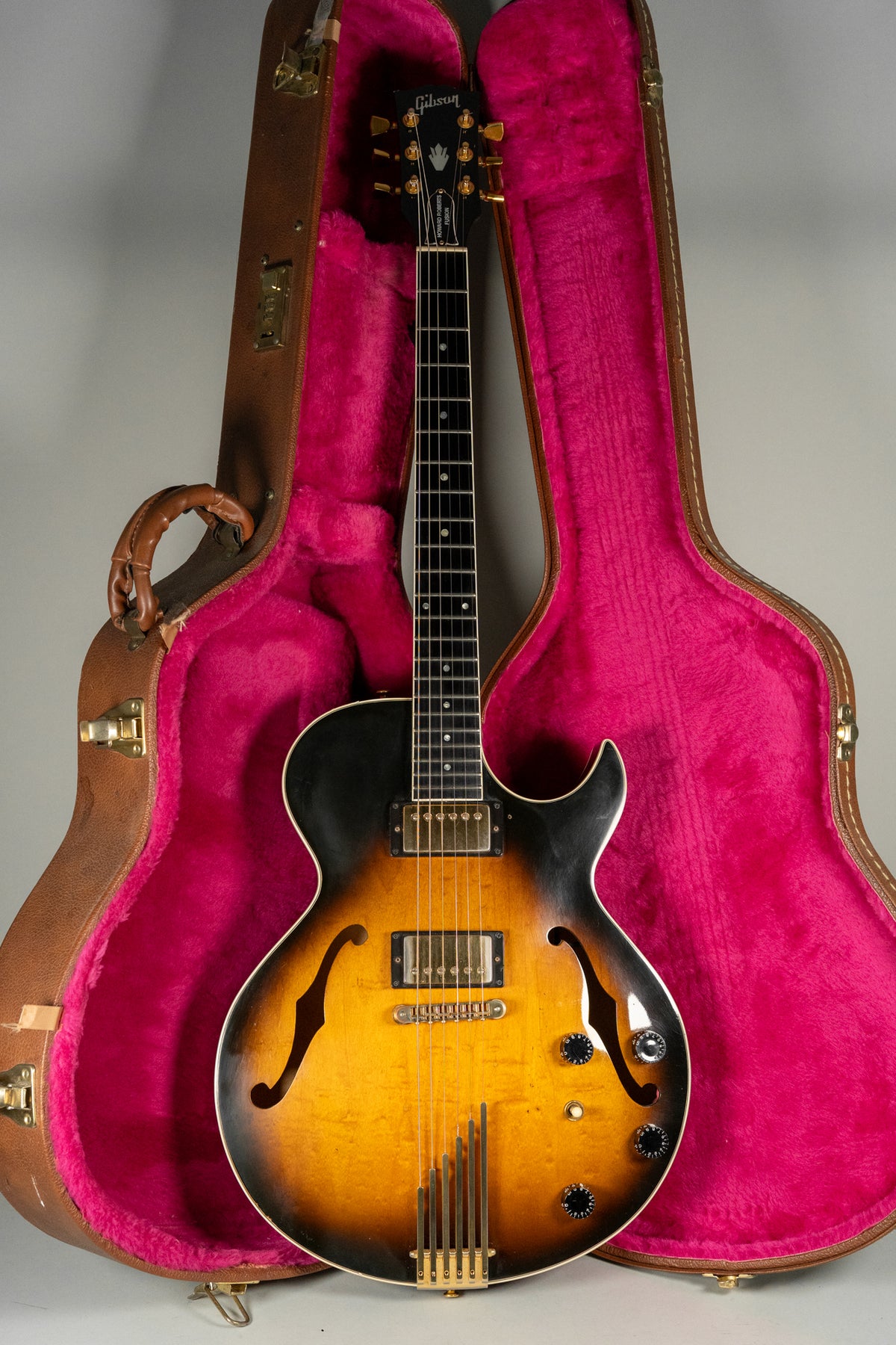 Full-length view of a sunburst archtop electric guitar with f-holes, gold hardware, and four black control knobs, resting in a pink-lined brown hard case.