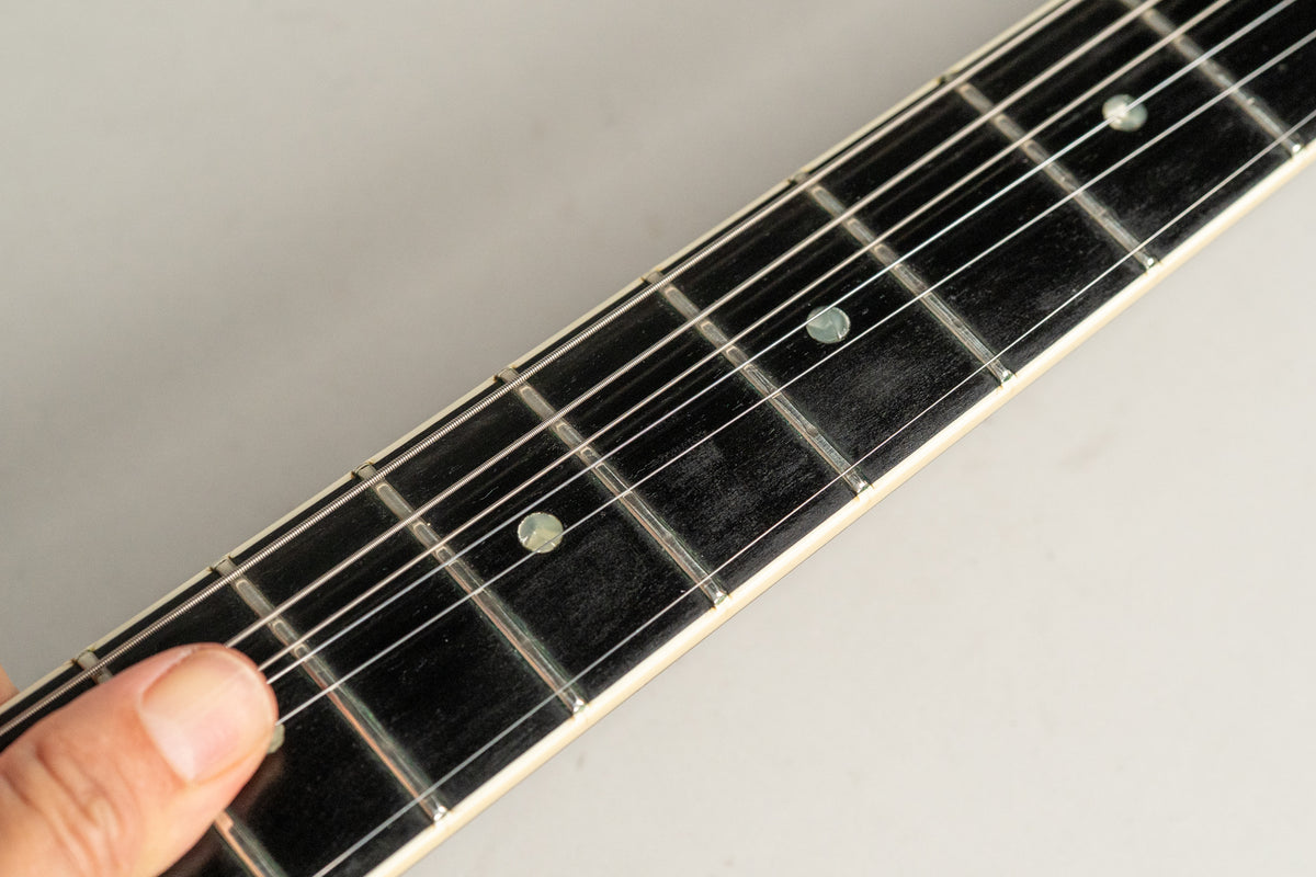 Close-up of the dark fretboard with light fret markers and strings, showing a finger pressing the first fret.